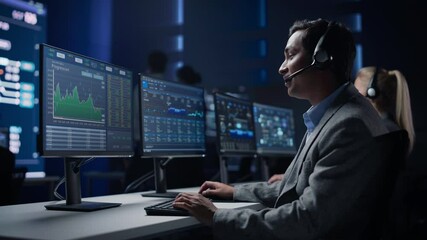 Confident Male Trader Talking into Headset Works on Computer with Screen Showing Finance Statistics, Charts Strategy, Stock. Big Stock Firm Monitoring Room with Team of Stock Market Specialists