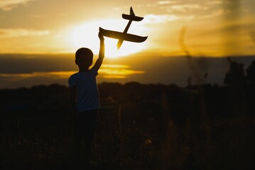 Happy child playing with a toy plane in nature during summer sunset. Boy in a white shirt with a plane in hands on wheat field. Kid holds a wooden airplane and dreams of being a pilot, on the nature