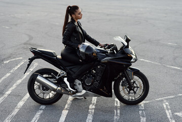 Attractive girl with long hair in black leather jacket and pants on outdoors parking with stylish sports motorcycle at sunset.