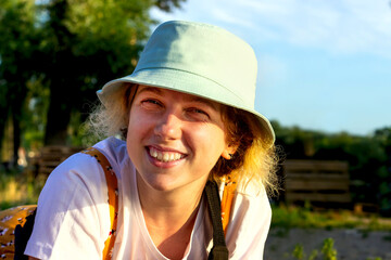 Close-up of a cute hipster girl sincerely smiling, laughing, looking at the camera. She's wearing a white t-shirt and a blue Panama hat. Summer outdoor recreation. Emotion of joy and happiness