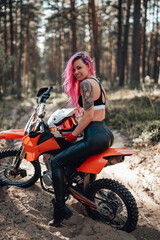 Fototapeta na wymiar Happy young woman with pink hair and tattoo on hand sitting on her modern motocross bike in woods