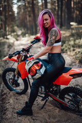 Naklejka na ściany i meble Beautiful young hipster girl with pink hair and tattoo on hand sitting on her modern motocross bike in woods, smiling and looking on the camera