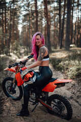 Obraz na płótnie Canvas Happy young woman with pink hair and tattoo on hand sitting on her modern motocross bike in woods