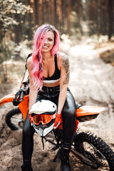Fototapeta na wymiar Beautiful young hipster girl with pink hair and tattoo on hand sitting on her modern motocross bike in woods, smiling and looking on the camera