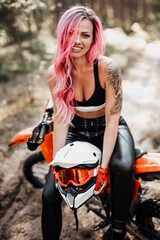Fototapeta na wymiar Beautiful young hipster girl with pink hair and tattoo on hand sitting on her modern motocross bike in woods, smiling and looking on the camera
