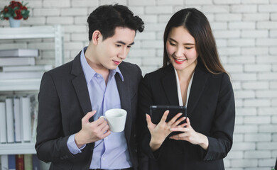 Happy successful of Young Asian businesswoman and businessman partners while working together with a tablet and business man holding a cup of coffee in office background,Working couple