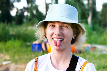 Cute funny hipster girl makes a face and shows her tongue. she's wearing a white t-shirt and a blue Panama hat. In nature against the background of green trees. Summer holidays and vacations