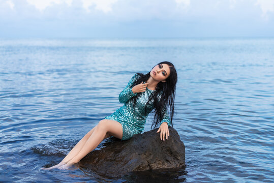  Young beautiful woman relaxing  on the big stone. Girl in a little green dress.Blue ocean.Girl traveler sits on the sea.fashion and  beauty concept