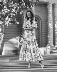 Elegant woman wearing summer fress, slty hat, sunglasses and holding salty handbag walking city street on a summer evening. Pretty beautiful caucasian girl with long brunette wavy hair