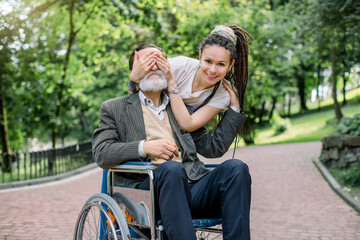 Obraz na płótnie Canvas Young smiling Caucasian woman with dreadlock hair covering the eyes of her senior disabled grandfather in wheelchair, making surprise for him on a walk in the park