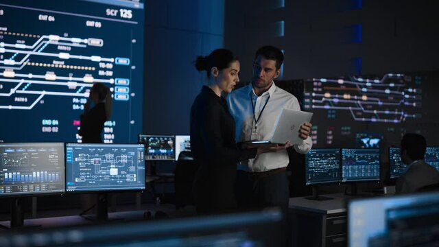 Project Leader and Chief Executive Discuss Data Shown on a Laptop. In Background Big Screen Showing Infographics of Infrastructure, Charts, System Analysis.Telecommunications Control Room