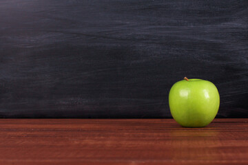 Single bright green apple on brown desk with blackboard texture in background. Copy space, close-up. School snacks or organic food concept
