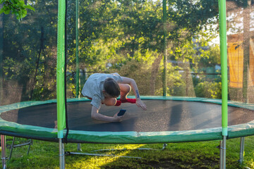 Cheerful eight years old boy having fun on trampoline – jumping and dancing with phone in hands....