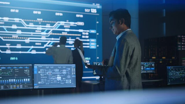Handsome Confident Male Project Manager works on a Laptop Standing in System Control Room. In Background Big Screen Showing Infographics of Infrastructure, Charts, System Analysis. Side View Arc Shot