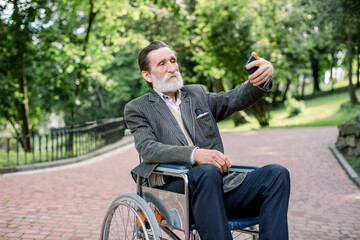 Happy senior disabled or handicapped bearded man sitting on a wheelchair in park, using his smartphone for photographing himself on the background of park landscape