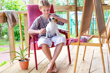 A Caucasian boy sits in a chair on the veranda and strokes the dog. Easel, painting, plein air. Portrait of a teenager with a dog. Aloe.