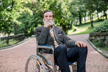 Thoughtful handsome senior bearded disabled man sitting in wheelchair walking in a city park. Concept of disabled people in everyday life