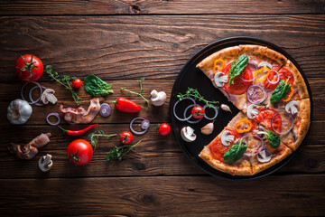 Pizza with bacon and ham without one slice on a slate black platter which is on wooden background, top view and copy space