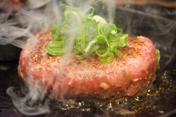 熱々の鉄板の上で湯気の立ち上る、まだ半生の調理中のハンバーグ、たっぷりの刻みネギ。一人前。外食、昼ご飯、食堂、オープンキッチン。口コミ、グルメ、行列、人気店。熱々、あつあつ。肉々しい。
