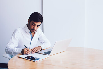 Concentrated male boss writing organisation plan in textbook sitting desktop with netbook at company office, serious businessman working with laptop device searching information for presentation