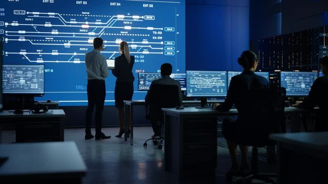 Project Leader and Chief Executive Discuss Data Shown on a Laptop. In Background Big Screen Showing Infographics of Infrastructure, Charts, System Analysis.Control Room Professionals Working
