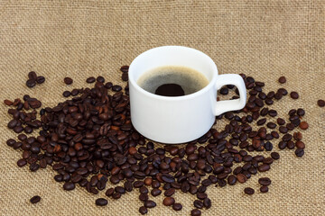 Hot espresso in ceramic cup on bagging, scattered coffee beans around cup.