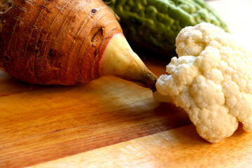 Taro root, cauliflower, and bitter melon on a wooden chopping board.