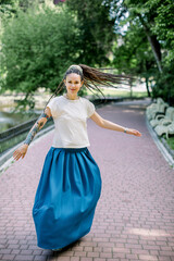 Fototapeta premium Charming young woman in white t-shirt and blue skirt, with dreadlocks hair style, walking and dancing in the park