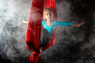 Fearless pretty little girl in a blue gymnastic suit shows a stunt aerial red ribbon surrounded by smoke on a black background. Concept of advanced acrobats gymnasts