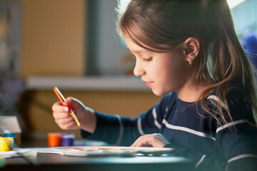 Side view of charming little caucasian smiling schoolgirl writes while sitting at table. Little cute girl draws picture. Concept of teaching children during self-isolation
