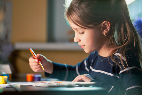 Side view of charming little caucasian smiling schoolgirl writes while sitting at table. Little cute girl draws picture. Concept of teaching children during self-isolation - Powered by Adobe