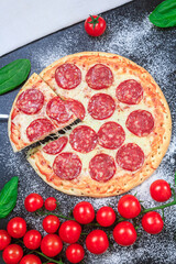 Pepperoni pizza one slice cut and rising cheese stretching golden dough, flour ripe cherry tomato branches with spinach on a black table. top view, flat lay, copy space, vertical orientation