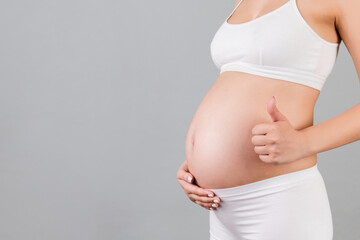 Cropped shot of positive pregnant woman in white underwear showing thumb up cool gesture over her baby bump at gray background. Happy maternity. Copy space