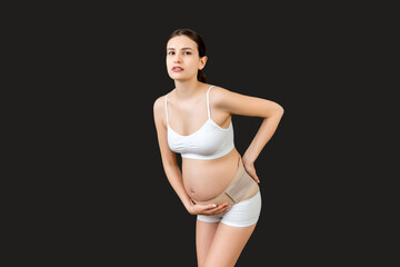 Portrait of supporting bandage against backache on pregnant woman in underwear at black background with copy space. Mother is suffering from pain in the back. Orthopedic abdominal support belt concept