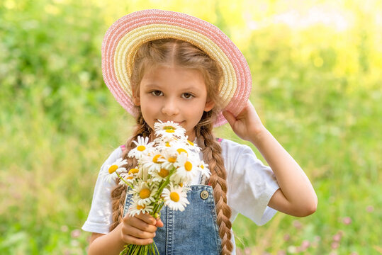 A little girl in a straw hat sniffs a bunch of wild flowers in a green clearing.