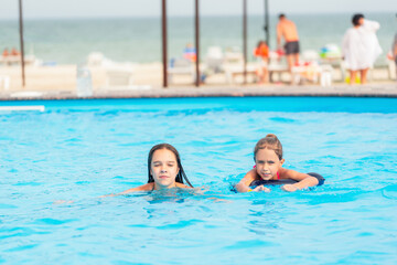 Fototapeta premium Two little sisters girls are swimming in a large pool with clear blue water of mine near the hotel on the background of the sea and the beach. Concept vacation tropical hot country with children