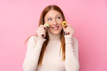 Young redhead woman over isolated pink background holding colorful French macarons and looking up