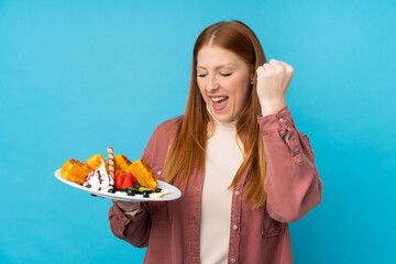 Young redhead woman holding waffles over isolated background celebrating a victory