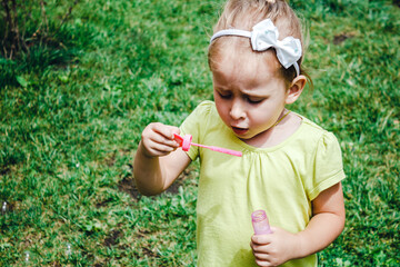 Summer outdoor activities for children. A little blonde girl in a yellow t-shirt blows soap bubbles against the background of green bright grass in the courtyard with a blur of focus.