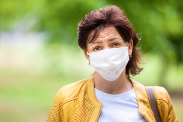 Portrait of sad unidentified woman posing in street in protective white medical mask on a sunny spring warm day. Concept of fatigue and sadness from coronovirus pandemic