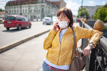 Fototapeta premium Funny middle-aged woman in a white protective mask talking on a smartphone while walking around the city on a warm spring day. Coronovirus pandemic and remote communication concept