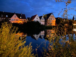 Häuserfront steht am Fluss, Häuser spiegeln sich im ruhigen Wasser. Auf der gegenüberliegenden...