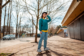 Naklejka na ściany i meble A young Caucasian hipster woman poses standing on a skateboard. In the background, an alley. Concept of sports lifestyle and street culture