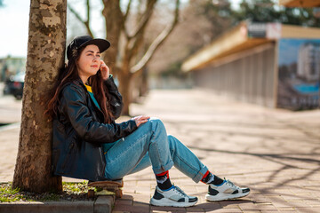 Portrait of a beautiful hipster teen girl sitting on a skateboard and looking into the distance. There's an alley with trees in the back. Copy space. Concept of sports lifestyle and street culture