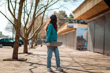 A young Caucasian hipster woman walks down the street with a skateboard in her hands and turns back. In the background, an alley. Concept of sports lifestyle and street culture © _KUBE_