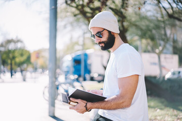 Young bearded talented male student dressed in casual clothes reading information from notepad while spending time at sunny street.Handsome smart hipster guy planning working schedule sitting outdoors