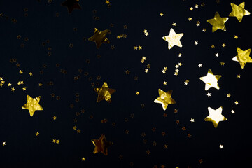 Fototapeta premium Black contrast color festive background with golden stars glitter confetti. Flat lay style with copy space for text, design. Trendy Holiday concept. Perfect backdrop for social media, blog, postcard