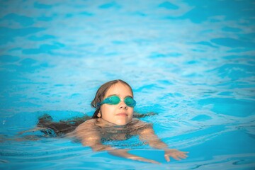 Charming girl teenager wearing waterproof glasses for the pool swims in the clear warm and blue water during the summer holidays. Concept of physical activity for children. Advertising space