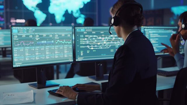Confident Female Data Scientist Works on Personal Computer Wearing a Headset in Big Infrastructure Control and Monitoring Room. Woman Engineer in a Call Center Office Room with Colleagues.