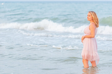 Fototapeta na wymiar Beautiful happy young pregnant woman in a summer light dress hugging her belly and walks along the sea waves on a sunny summer evening during vacation. Natural Parenthood Concept. Copyspace